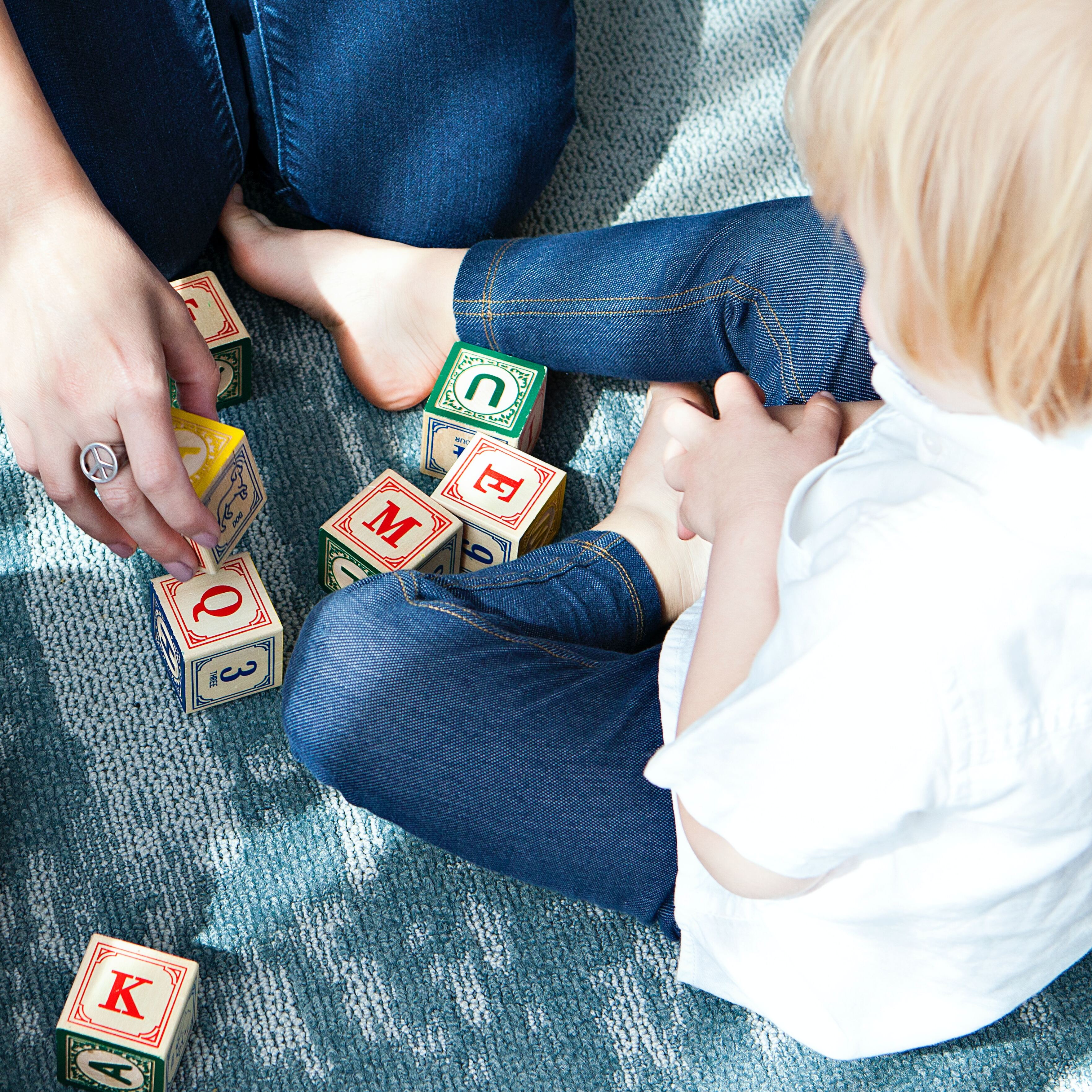 Ein Kind im Kindergartenalter mit dem Rücken zum Betrachter sitzend, vor dem Kind eine Hand, die einen Hölzwürfel greift. 