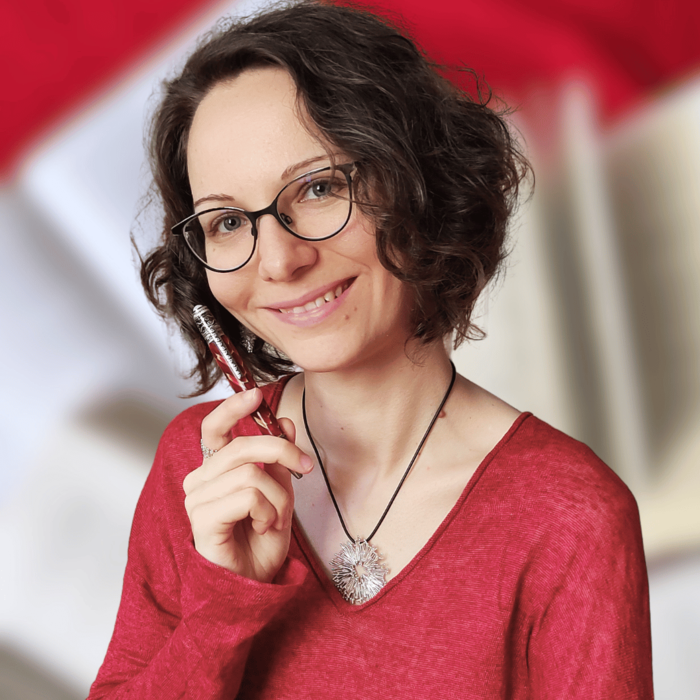 Frau mit Brille und braunen Locken hält einen Stift in der Hand und lächelt.